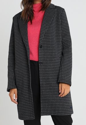 Femme portant un long manteau pied-de-poule noir et gris par-dessus un col roulé rose vif et un pantalon noir, debout devant un fond uni.