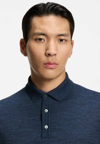 Mörkblå polo-skjorta i texturerat tyg med krage, tre knappar och ett subtilt mönster. Fokusera på nacke och övre bröstparti.