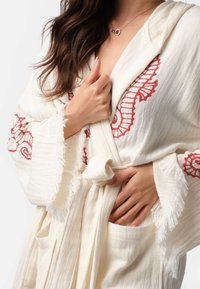 Biely plášť s červenými vyšívanými vzormi morskej koníka, otvorený predný diel, zapínanie na opasok, strapcové okraje a dve bočné vrecká. Textúrovaná tkanina.