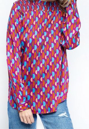 Blusa a maniche lunghe di un viola vivace con motivo geometrico multicolore. Presenta un colletto smock e polsini con bottoni, abbinata a jeans strappati.