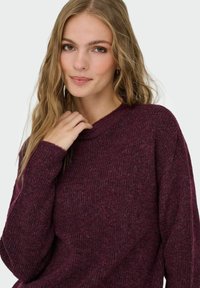 Burgunderfarbener Rippstrick-Pullover mit rundem Halsausschnitt. Weicher Textur und lässiger Schnitt. Das Model berührt leicht den Pullover in der Schulterregion.
