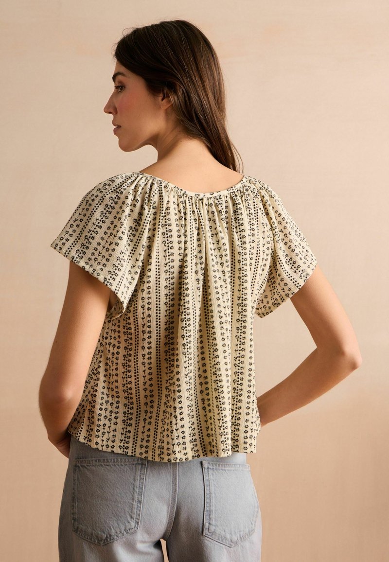 Blouse en mélange de coton avec un fond crème et des rayures florales noires. Présente une coupe ample et des manches courtes plissées.