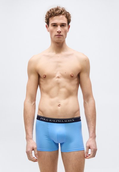 Νέος άνδρας που στέκεται, φοράει ανοιχτό γαλάζιο boxer briefs Polo Ralph Lauren με μαύρη μέση, μπροστά σε απλό ανοιχτό γκρι φόντο.