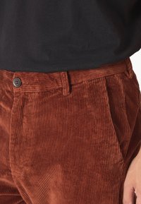 Pantaloni od kordura u boji hrđe s vertikalnom rebrastom teksturom, s jednim prednjim gumbom i standardnim dizajnom struka.