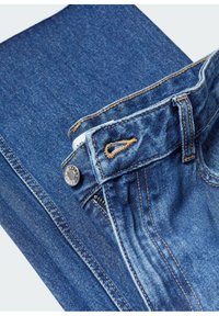 Gros plan sur un jean en denim bleu montrant un bouton, une fermeture éclair, une passoire de ceinture et des coutures détaillées sur le tissu.