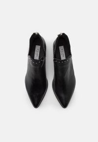 Une paire de bottes de cheville noires en cuir à bout pointu avec des clous argentés et une fermeture éclair à l'arrière, vue d'en haut sur un fond blanc.
