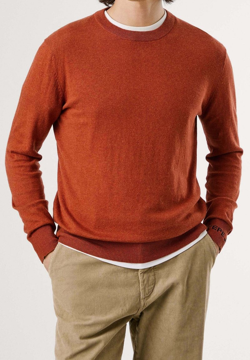Homme portant un pull à manches longues orange brûlé sur une chemise blanche, associé à un pantalon en velours côtelé beige, mains dans les poches, debout devant un fond uni.