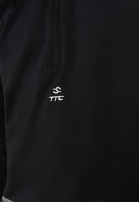 Svart atletisk topp med en kvart glidelås i kragen. Har en liten hvit brodert logo på brystet. Glatt, elastisk stoff.