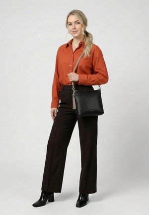 Frau mit blonden Haaren, die ein oranges Hemd, schwarze Hose, schwarze Stiefel trägt und eine schwarze Umhängetasche mit beigem Riemen bei sich hat.