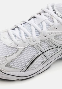 Hvit atletisk sko med mesh-overdel med heksagonale mønstre, syntetiske lærdetaljer, hvite snører og en svart gummisåle.