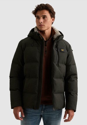 Homme portant une veste matelassée vert foncé avec capuche et un pull zippé marron, debout devant un fond uni clair.