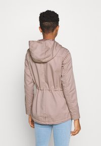 Person with short curly hair wearing a light brown hooded jacket and light blue jeans, facing away against a plain white background.