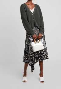Khakifarbener gerippter Cardigan mit Knöpfen, übereinander getragen über einem fließenden Rock mit schwarz-weißen Tiermotiven. Klare Handtasche mit weißem Innenbereich und Holzgriffen.