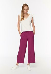 Witte mouwloze top met V-hals en zijstrikjes, gecombineerd met brede, elastische taillebroek in bessen kleur, gedragen met witte sneakers.