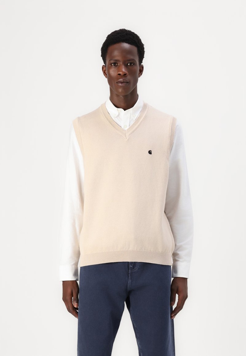 Μπεζ πλεκτό γιλέκο με V-λαιμό, φορεμένο πάνω από ένα λευκό πουκάμισο με γιακά. Διαθέτει ένα μικρό σκούρο λογότυπο στο αριστερό στήθος. Κανονική εφαρμογή.