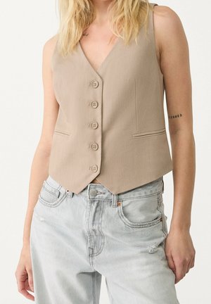 Woman wearing a beige buttoned vest and light gray distressed denim jeans with her hand resting by her side.