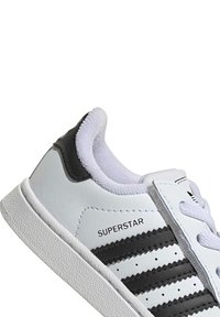 Baltas sporta apavu ar melniem trim svītrām un zīmolu. Iezīmes: gluda sintētiska virsma, strukturēta zole un mīkstināta apkakle komfortam.