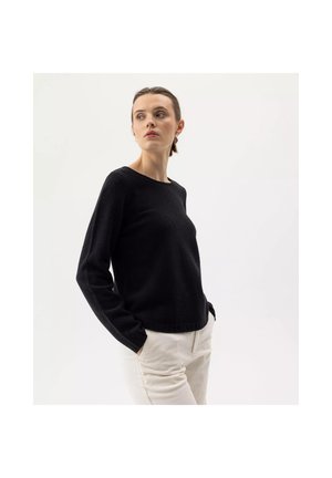 Vrouw met donker haar in een zwarte trui met lange mouwen en lichtgekleurde broek poseert met handen in de zakken, kijkend naar de zijkant tegen een effen achtergrond.