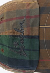 Brūns pleds cepure, kas izgatavota no auduma ar zaļām, sarkanām un dzeltenām akcentiem. Satur izšūtu logotipu tumšās diegās un pogu detaļu augšpusē.