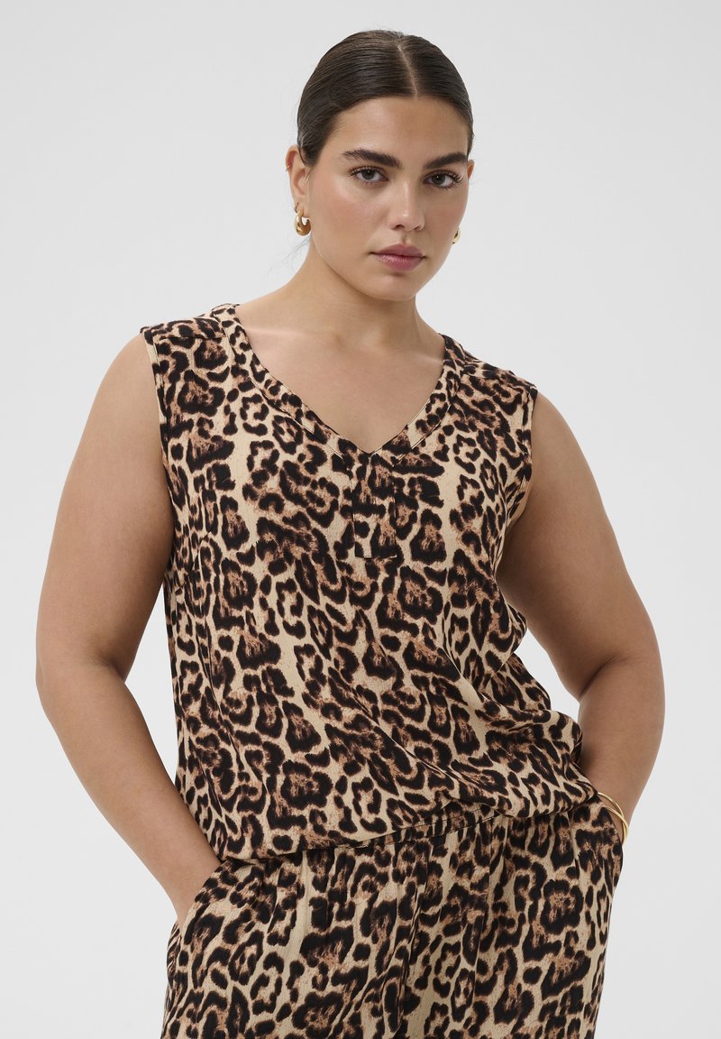 Femme aux cheveux foncés attachés, portant des boucles d'oreilles créoles dorées et un haut sans manches imprimé léopard avec un pantalon assorti, mains dans les poches.