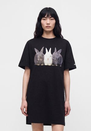 Femme portant une robe T-shirt noire oversize avec trois lapins réalistes — deux gris et un blanc — imprimés sur la poitrine.