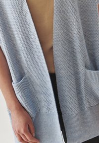 Világoskék kötött cardigan, textúrált szövéssel, nyitott elöl, és két elülső zsebbel. Bordás mandzsettákkal és aljjal rendelkezik.