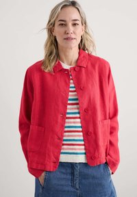 Giacca di lino rossa con colletto, bottoni frontali e due tasche. Indossata sopra un maglione a righe blu, rosso e bianco. Design semplice e rilassato.
