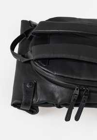 Gefalteter schwarzer Rucksack aus Leder und Stoff mit gepolsterten Trägern und zwei Reißverschlusslaschen an der Vorderseite, auf weißem Hintergrund.