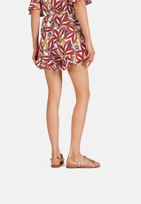 Romper con motivo tropical in rosso, arancione, verde e nero; modello che indossa sandali metallici oro rosa con decorazioni.