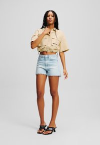 Camicetta beige a maniche corte con bottoni, con due tasche frontali, abbinata a pantaloni corti in denim chiaro con bordi sfilacciati e sandali neri con logo.