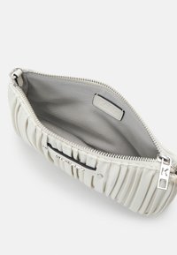 Pochette blanche plissée avec fermeture éclair argentée partiellement ouverte, montrant l'intérieur en tissu gris lisse avec une petite étiquette à l'intérieur.