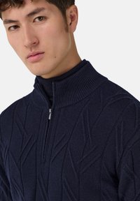 Mladý muž s krátkými tmavými vlasy, oblečený v námořnickou modrou pletenou svetr s texturou, se zipem do poloviny a vysokým žebrovaným límcem.