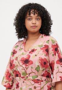 Floral dress in pink with a v-neckline, featuring large red and green flower patterns, and loose short sleeves. Smooth texture.