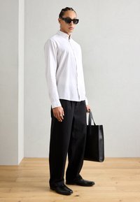 Camisa blanca de botones, pantalones anchos negros, zapatos negros y gafas de sol. Sosteniendo una bolsa de tote negra. Tejido suave, diseño minimalista.