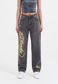 Grå denimbukser med løs pasform, der har gul "Ed Hardy" tekst og et lille farverigt grafisk motiv nær kanten, båret med hvide sneakers.