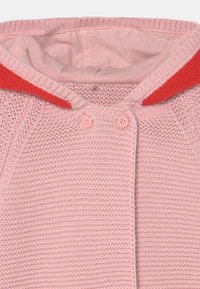 Růžový pletený cardigan se solidním červeným límečkem, dvěma růžovými knoflíky a měkkou vnitřní podšívkou. Má texturovaný, otevřený pletený vzor.