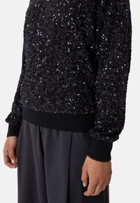 Maglione nero con paillettes, caratterizzato da una superficie testurizzata, polsini e orlo a costine. Abbinato a pantaloni scuri, mostra una vista ravvicinata del tessuto.