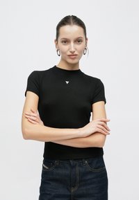 T-shirt nera aderente a maniche corte con collo alto e piccolo logo triangolare sul petto, realizzata in tessuto liscio, abbinata a jeans scuri.