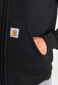 Sudadera negra con un bolsillo frontal, puños y dobladillo de canalé. Parche del logo en naranja y blanco en la zona izquierda del bolsillo. Textura suave.