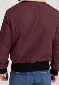 Mężczyzna ubrany w bordo bomberkę z czarnymi mankietami i paskiem w pasie, w połączeniu z niebieskimi dżinsami, widoczny od tyłu.