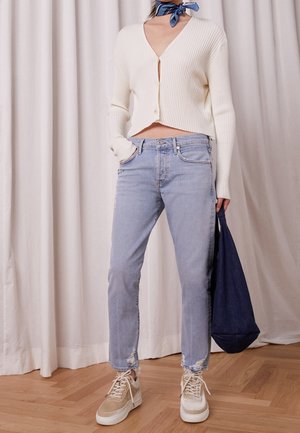 Person iført hvid, ribbet cropped cardigan, lyseblå slidte jeans, beige sneakers, blå halsedisse, holder navy mulepose indendørs.