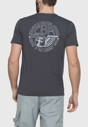 Uomo che indossa una t-shirt grigio scuro con un logo bianco circolare che mostra un'aquila e del testo sul retro, abbinata a pantaloncini cargo grigio chiaro.