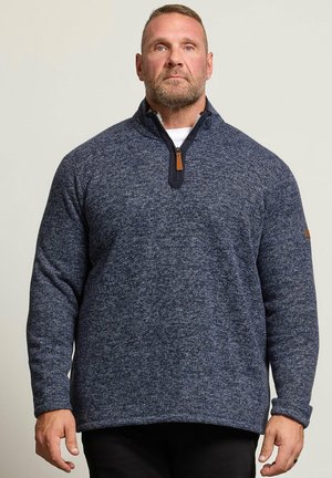 Homme avec une barbe courte portant un pull texturé bleu marine à fermeture éclair quart et un pantalon noir, debout devant un fond uni.