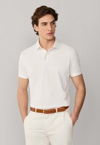 Homme portant un polo à manches courtes blanc, un pantalon blanc et une ceinture en cuir marron, debout les mains dans les poches.