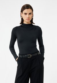 Nainen, jolla on tummat hiukset, käyttää kultaisia renkaankorviksia, tiukkaa mustaa pitkähihaista paitaa, mustaa vyötä ja löysiä mustia housuja, kädet taskuissa.