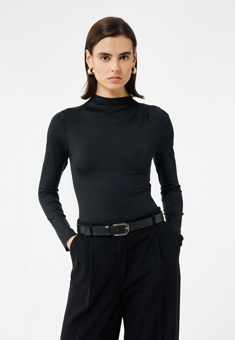 Nainen, jolla on tummat hiukset, käyttää kultaisia renkaankorviksia, tiukkaa mustaa pitkähihaista paitaa, mustaa vyötä ja löysiä mustia housuja, kädet taskuissa.