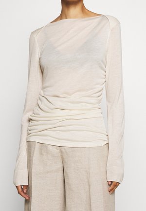 Long-sleeve cream top with a boat neckline, featuring soft, draped fabric and ribbed cuffs. Styled with loose beige trousers.