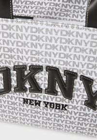 Balta drobės pirkinių krepšys, puošiamas pasikartojančiu DKNY logotipo raštu, su dideliais juodais užrašais ir juodais odiniais akcentais.