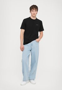 Juoda trumpomis rankovėmis marškinėliai su EA7 logotipu, derinami su šviesiai mėlynais plačiomis kelnėmis ir baltais sportbačiais, stovint priešais vientisą foną.