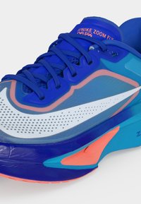 Μπλε αθλητικό παπούτσι Nike Zoom Fly με λευκό σήμα, κοραλλί και τιρκουάζ λεπτομέρειες και μπλε κορδόνια σε λευκό φόντο.